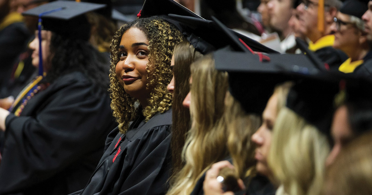 Graduate looks toward the camera while seated among other commencement participants.