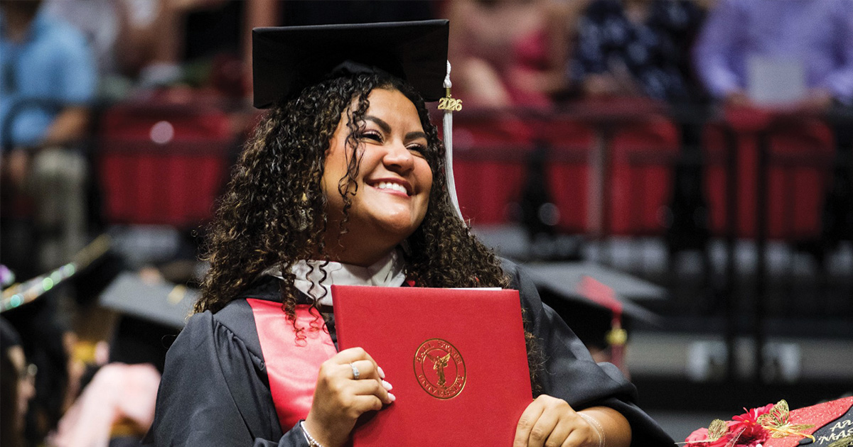 Graduate smiles while holding a red Ball State diploma cover.