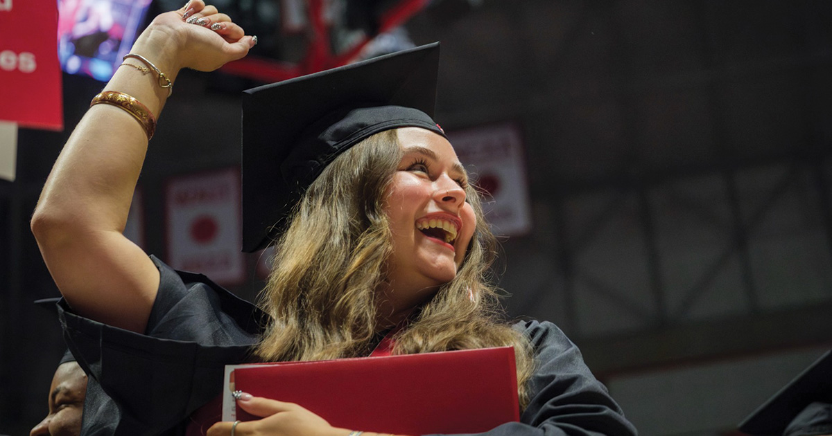 Graduate raises her arm and celebrates while holding a red diploma cover.