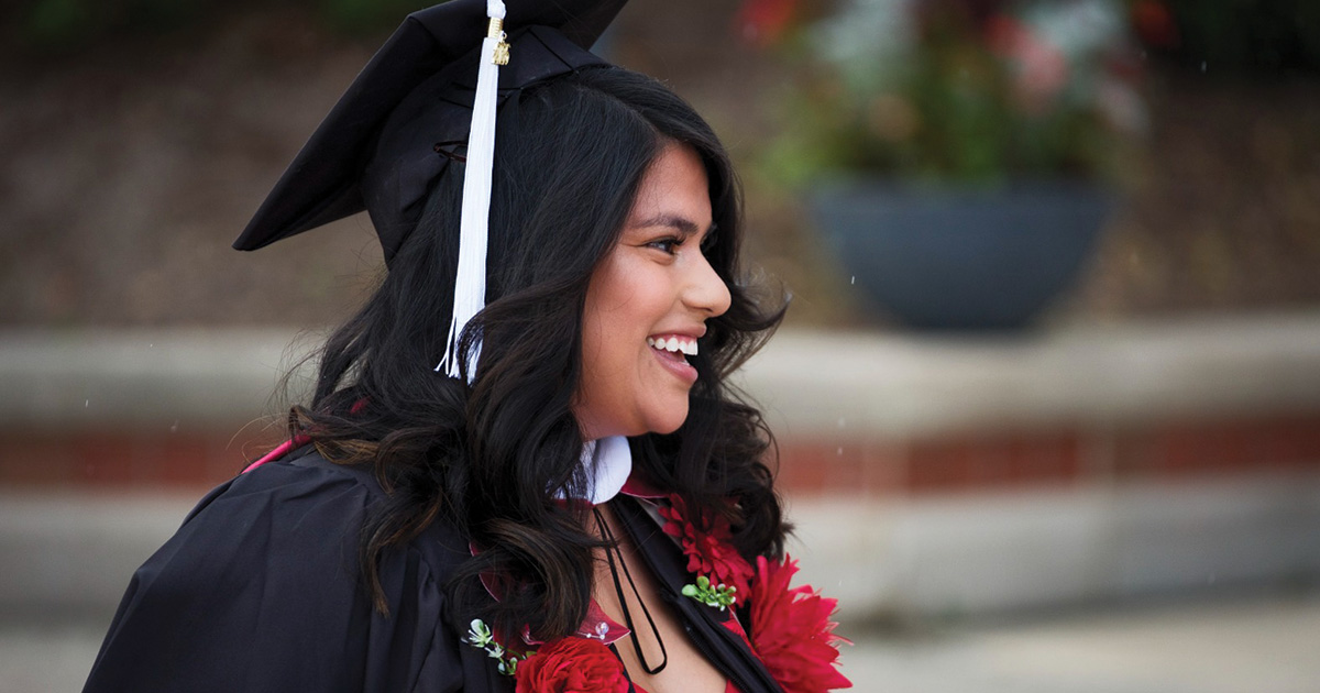 Graduate smiles during an outdoor commencement celebration, wearing red flowers over her gown.