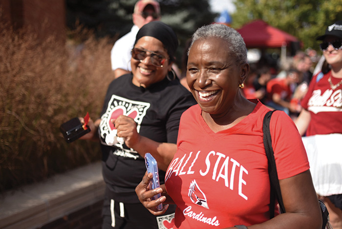 Two smiling Ball State supporters walk outdoors during a campus event, wearing Ball State apparel.
